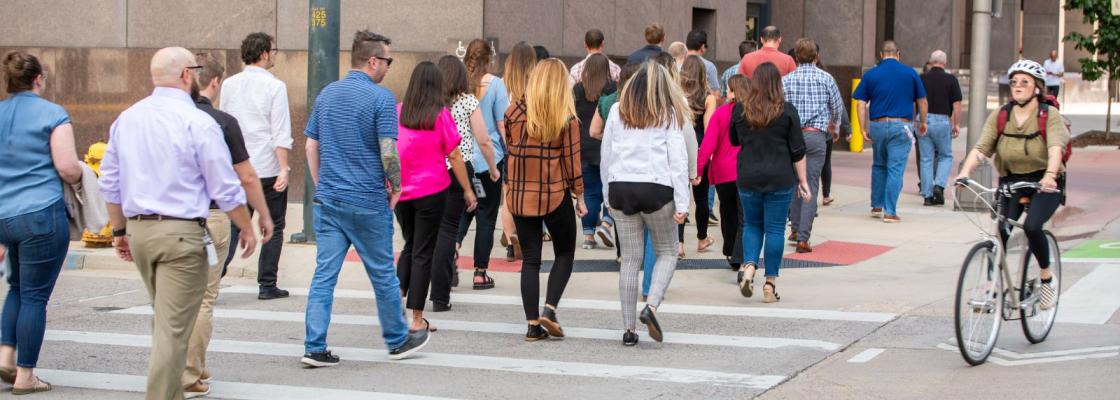 A large group of commuters crossing a crosswalk with a person riding a bike in the bike lane traveling the opposite direction.