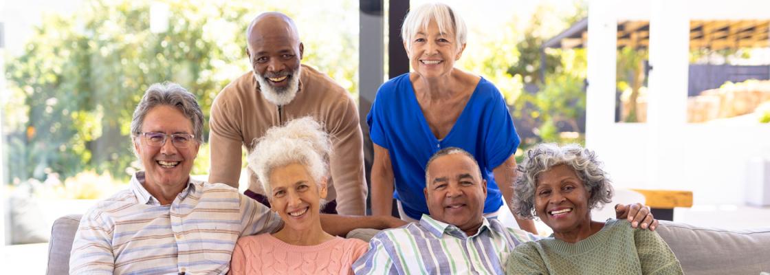 A racially diverse group of four older adults sit on a couch from left to right of man, woman, man woman. Two older adults stand behind the couch in the order of man and woman.