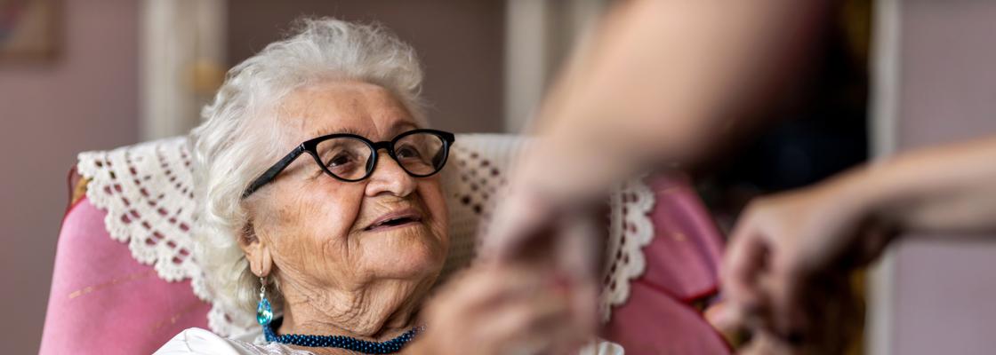 An older adult woman sits in a padded chair grasp hands of a person whose face is not pictured.