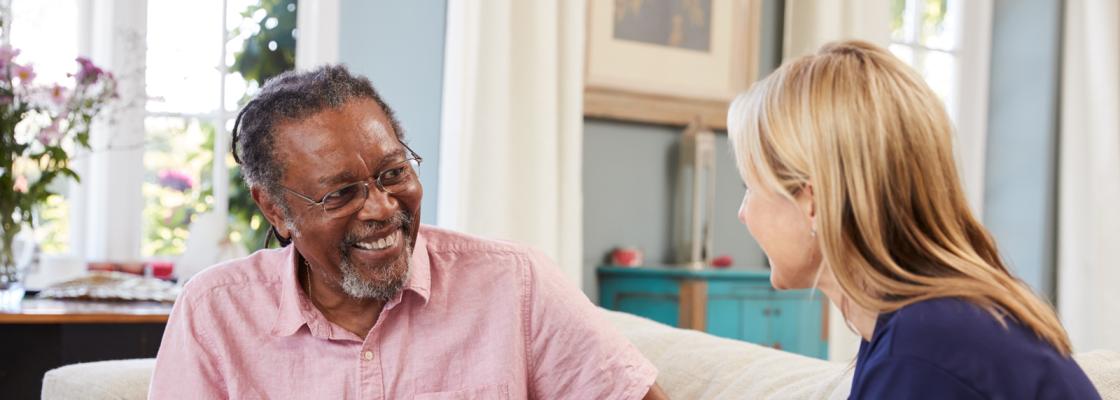 An older adult Black man sits on a sofa with an aide who is holding a clipboard with forms.
