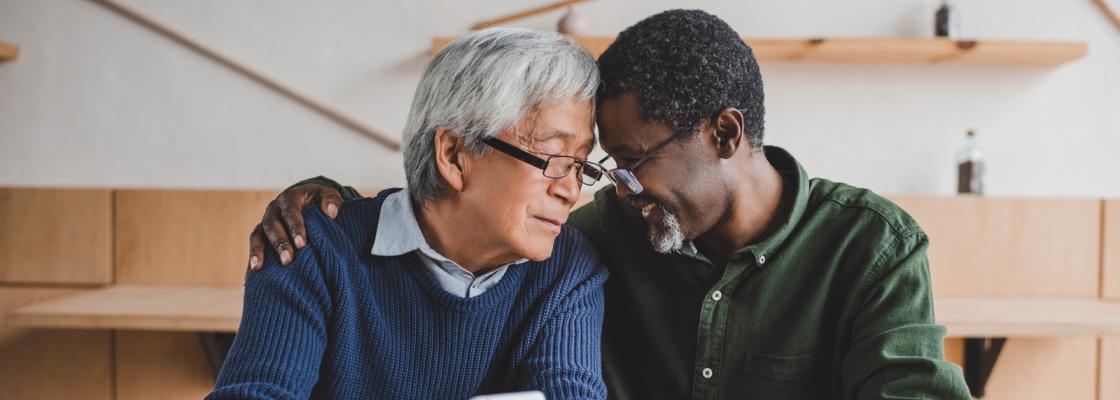 An older adult man of Asian descent and an older adult man of Black descent embrace each other.