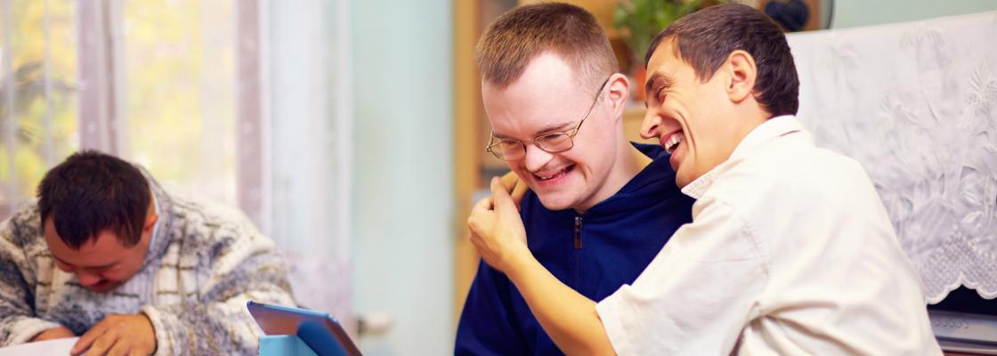 Two men with disabilities are sitting at a table laughing at the images displayed on a tablet electronic device.