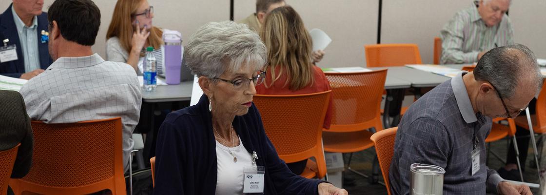 Several people sitting at two long conference tables and looking at printed materials. 