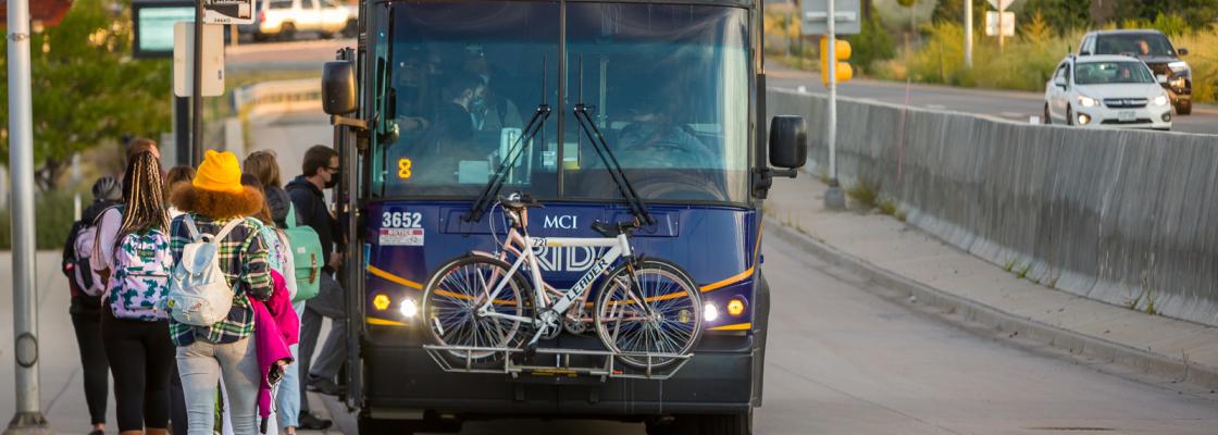 group of people waiting to board a bus that has two bikes in a bike rack on the front.