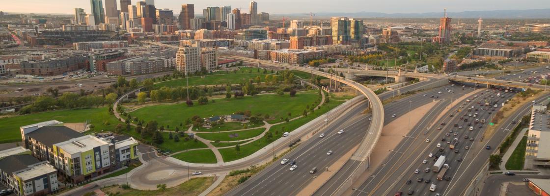 ars on a highway with a park and the Denver city skyline in the background.