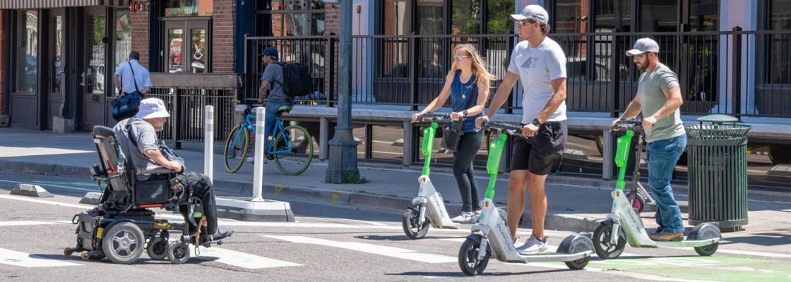 Three people on scooters and one person in a wheelchair crossing a crosswalk on a city street.