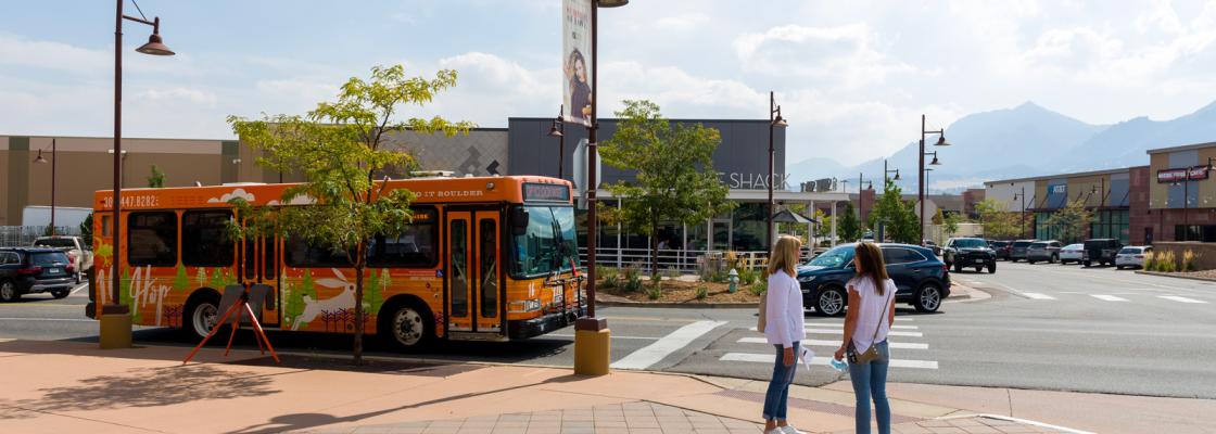 Two people standing on a sidewalk in a commercial district as a bus and car pass by on the street next to them.