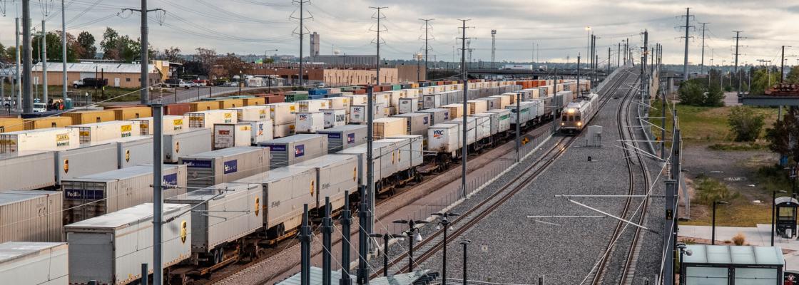 A commuter rail train pulling into a station with several freight train cars sitting on the tracks next to it.