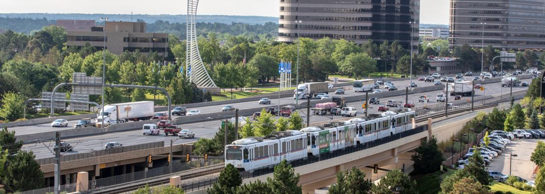 Cars and trucks moving along a highway next to a light rail train on elevated tracks, with green trees and two tall buildings in the background.