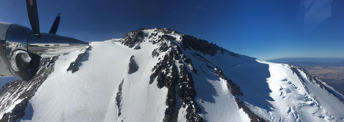 A snow-covered mountain peak above treeline on cloudless day, with an airplane propeller in the foreground.