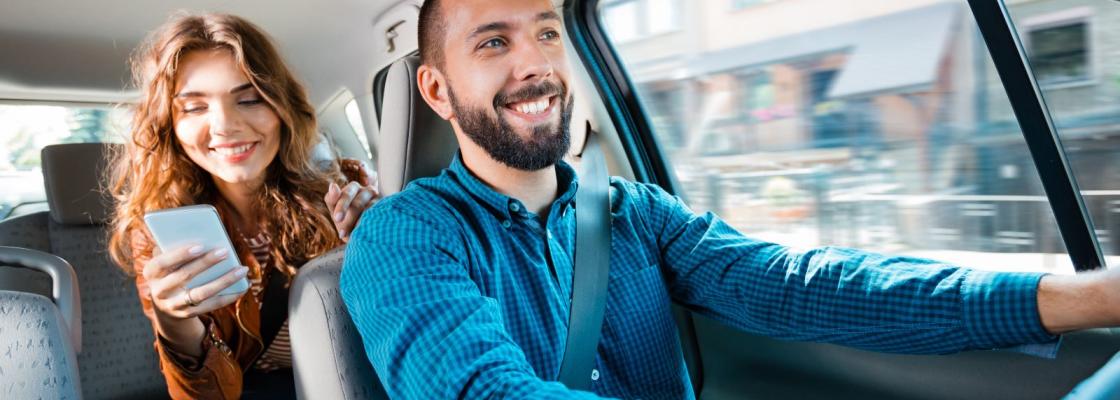 A person looking at their phone in the backseat of a car with a person directly in front of them in the driver's seat driving the car.