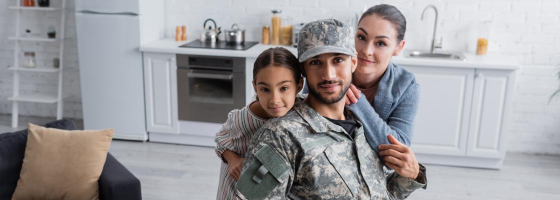 Dressed in military uniform, a soldier sits in a wheelchair while being embraced by his wife and daughter.
