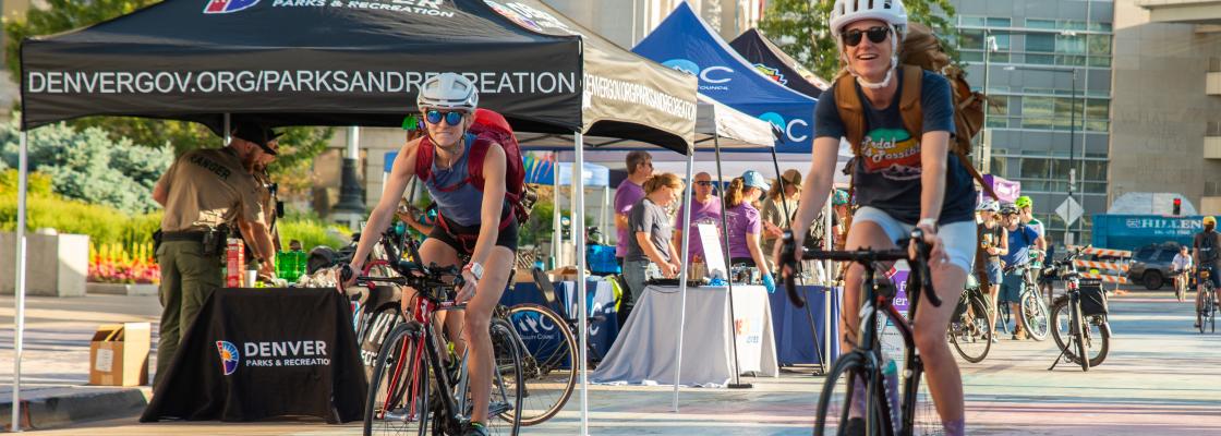 Two people riding bicycles with a crowd of people and booths set up in the background. This image was taken on Bike to Work Day 2024.