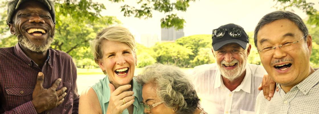 Four older adults,  a Black man, a white woman, an Asian woman, a white man and an Asian man are standing while laughing in a park setting.