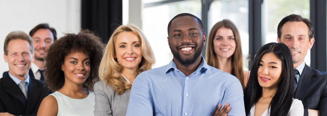 Several people of various races and ethnicities, all wearing business attire, stand smiling in front of a large office window.