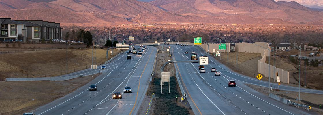 Several cars travel along highway C470 at sunrise, with the Front Range foothills in the background.