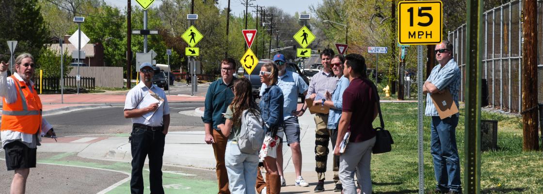 A group of people at a road crosswalk listening to a man in an orange vest talking. The people in the group have clipboards in their hands and are taking notes.