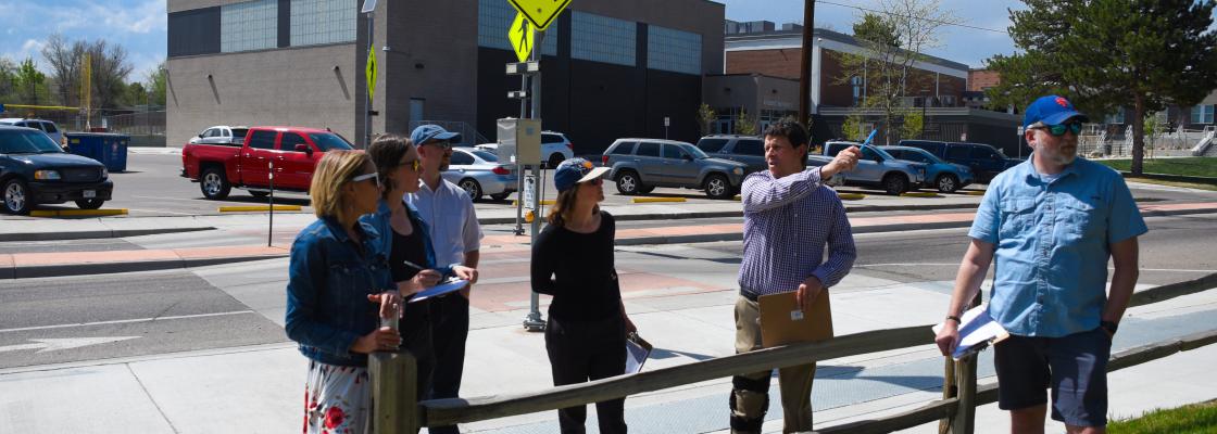 A group of six people standing on a sidewalk in front of a school, while the man holding a clipboard points to the left.
