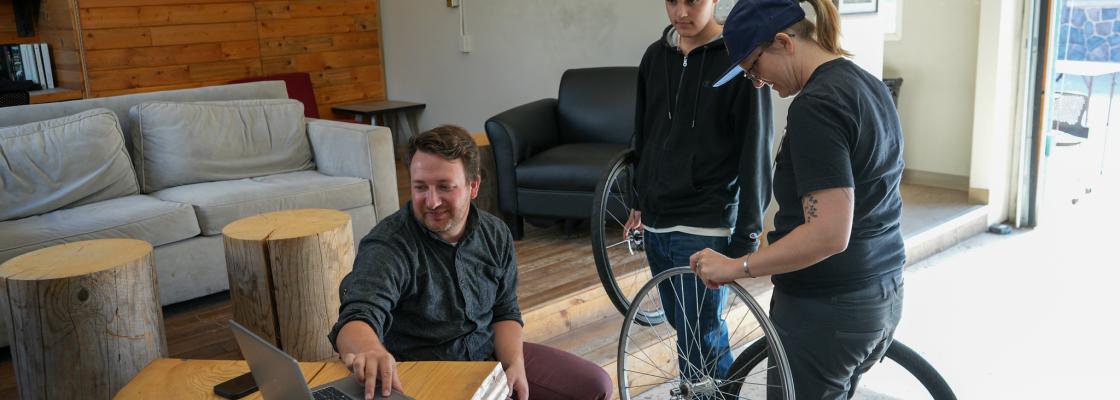 A man sitting at a table with a labtop while two people are stand holding bicycle parts.
