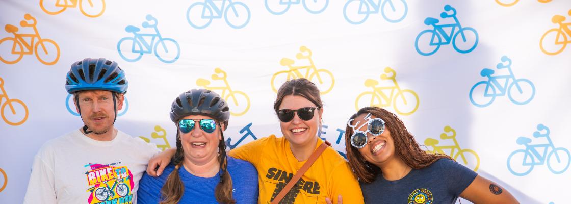 Four people smile in front of a Bike to Work Day backdrop in t-shirts, shorts, sunglasses and bicycle helmets.