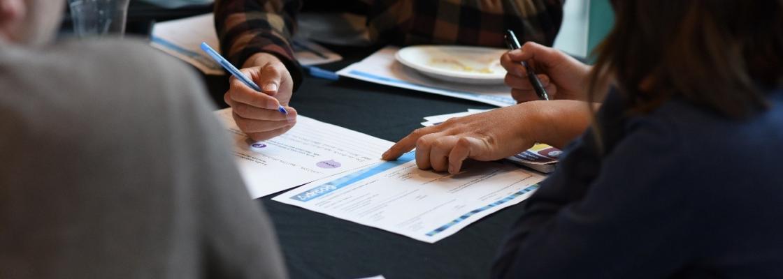 Cuatro personas sentadas alrededor de una mesa con bolígrafos en las manos, revisando documentos distribuidos sobre la mesa. 