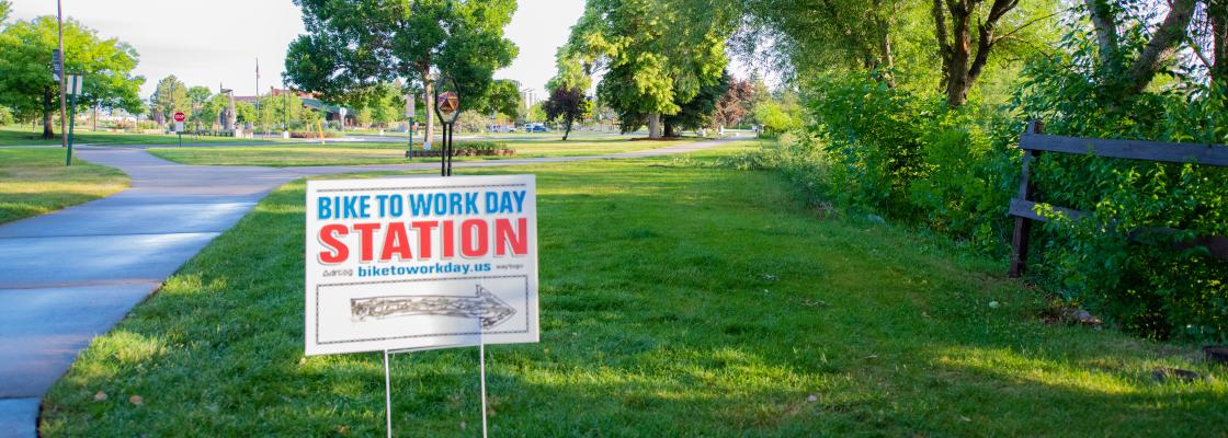 A wayfinding sign is staked along a trail and reads, "Bike to Work Day Station," with a hand drawn arrow indicating that cyclists turn to the right. The sign is along a paved sidewalk path lined with grass and trees.