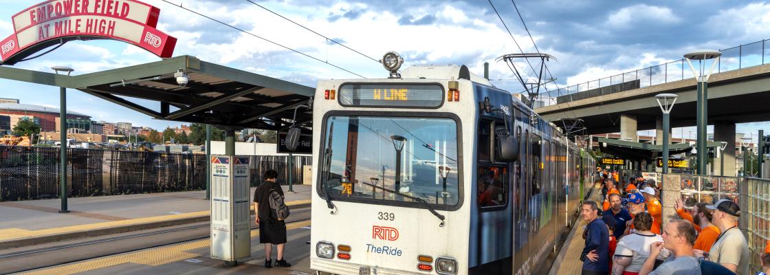 The image shows a light rail train at the "Empower Field at Mile High" station. A crowd of people is waiting on the platform as the train marked "W Line" is parked on the tracks. The scene takes place under a partly cloudy sky.